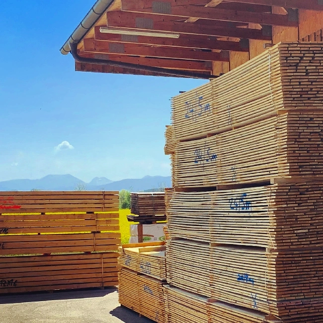 Bild zeigt mehrere Holzstappel mit besäumten Brettern vor schönem Hintergrund mit Bergen und blauem Himmel.